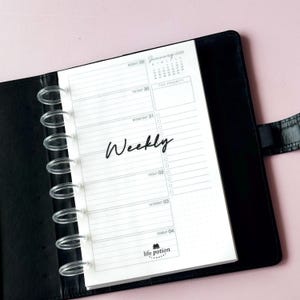 May include: A black disc-bound planner with a white weekly insert page. The page features the text Weekly, January 2026, and Life Potion Planners. It includes sections for days of the week, a top priority box, and a small monthly calendar grid.