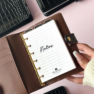 May include: Brown leather planner with gold-colored rings, open to a lined notepad with the word "Notes" in black script. The notepad has labeled tabs for "monthly", "weekly", and "password". The planner is on a pink surface.