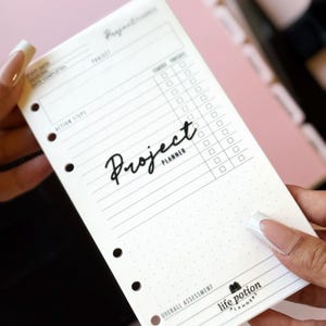 May include: A white paper project planner insert with black text and lines. The page features a project title area, action steps with checkboxes, a dot grid section, and the text Project Planner and Life Potion Planners at the bottom.