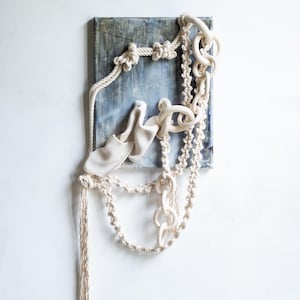 May include: Abstract wall art featuring a textured blue and gray canvas with cream-colored rope and chain elements. The design includes knotted rope, a draped fabric piece, and a chain with circular links, creating a unique, tactile composition.