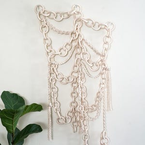 May include: Off-white wall hanging featuring a chain design. The artwork is composed of large, interlocking chain links, with smaller chains and knotted rope accents. Tassels hang from the bottom. A houseplant is visible in the lower left corner.