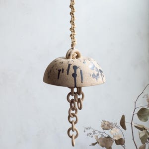 May include: A beige ceramic wind chime with black paint splatters, suspended by a knotted rope. The chime features a bell-shaped top and a chain of interlocking rings. Decorative and rustic.