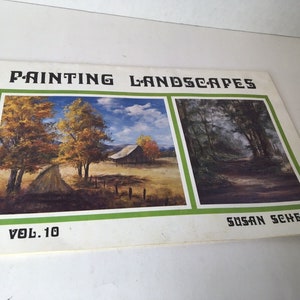 May include: A book titled "Painting Landscapes" by Susan Scheewe, volume 10, with two landscape paintings on the cover. The left painting shows a country road with trees and a barn in the distance. The right painting shows a forest path with trees on both sides.