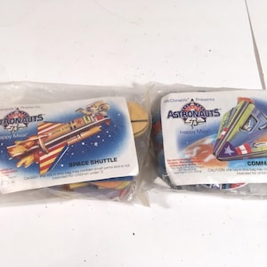 Könnte beinhalten: Zwei versiegelte McDonald's Happy Meal-Tüten mit Spielzeug zum Thema Astronauten. Eine Tüte zeigt eine Space Shuttle-Grafik mit roten, weißen und blauen Akzenten. Die andere zeigt ein Command Module.