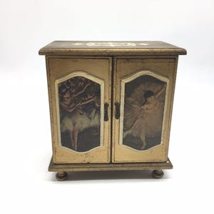 May include: A small, vintage music box with a gold-painted finish. The box features two doors with framed images of ballerinas. The top of the box has a decorative design. The box sits on four small, rounded feet.