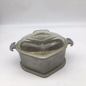 May include: A heart-shaped aluminum casserole dish with a textured matte finish and two side handles. The dish includes a clear, domed glass lid featuring a geometric diamond pattern and a central handle, providing a secure fit for the base.