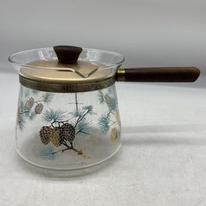 Könnte beinhalten: Ein klarer Glas-Kochtopf mit Holzgriff und Deckel. Das Glas ist mit einem Kiefernzapfen- und Zweigdesign in Gold-, Türkis- und Brauntönen verziert. Der Deckel ist aus goldfarbenem Metall mit einem dunkelbraunen Holzknopf.
