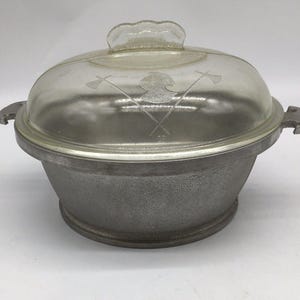 May include: A round, textured gray metal cooking pot with two side handles and a clear glass dome lid. The lid features an etched design of a profile silhouette of a person wearing a helmet, positioned between two crossed axes, with a handle on top.