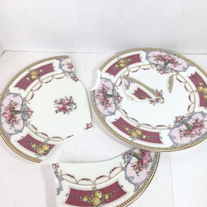 May include: A broken white porcelain plate with a pink and gold floral design. The plate is broken into three pieces.