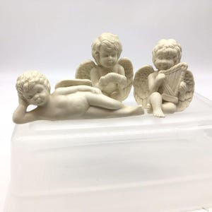 May include: Three off-white cherub figurines. One cherub is reclining, another is seated and holding a musical instrument, and the third is seated and holding a musical instrument. The cherubs have wings and curly hair.