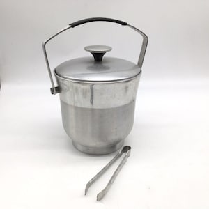 Puede incluir: Una cubitera de aluminio cepillado con tapa a juego que incluye un pomo negro. El recipiente cuenta con un asa abatible con agarre negro y viene con un par de pinzas para hielo de metal a juego situadas delante del contenedor.