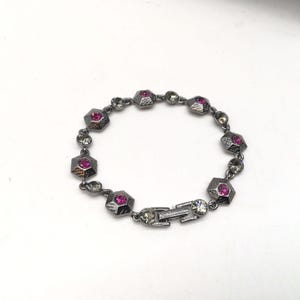 Puede incluir: Un brazalete plateado con un diseño de cadena y piedras preciosas rosas. El brazalete tiene un cierre de broche.