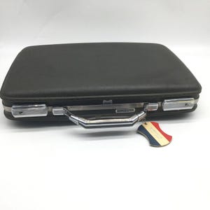 Könnte beinhalten: Ein dunkelgrauer Vintage-Koffer mit silberner Hardware, einem Griff und einem Anhänger. Der Koffer hat eine rechteckige Form und eine strukturierte Oberfläche. Der Anhänger ist rot, weiß und blau.
