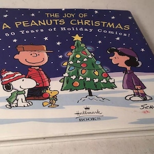 May include: A hardcover book titled "The Joy of a Peanuts Christmas" with Snoopy, Charlie Brown, and Lucy standing near a decorated Christmas tree. The cover is dark blue with white stars and the Hallmark Books logo.