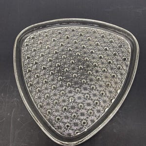 May include: Clear glass triangular dish with a detailed, textured pattern. The dish features a repeating design of small circles and starburst shapes, creating a decorative surface. The edges are slightly rounded, and the dish is set against a dark background.