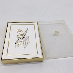 Peut inclure: Un ensemble de cartes de correspondance rectangulaires rangées dans une boîte dorée avec un couvercle en plastique transparent. Chaque carte présente une illustration centrée de trois coquillages aux motifs bruns et blancs.