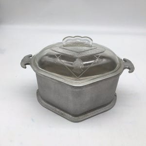 Puede incluir: Una cacerola de metal de forma cuadrada con un acabado gris texturizado y dos asas laterales. El recipiente incluye una tapa de vidrio transparente con un diseño central grabado en forma de diamante que muestra un pequeño retrato de perfil y flechas cruzadas.