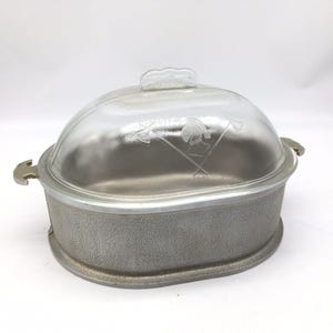 May include: An oval-shaped metal roaster with a textured silver-tone exterior and two side handles. The roaster includes a clear glass dome lid featuring an etched design of a helmet positioned between two crossed axes, topped with a glass handle.
