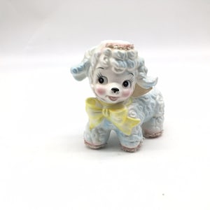 May include: A ceramic lamb figurine featuring a white body with blue accents, a yellow bow around its neck, and pink blush on its cheeks. The lamb has large dark eyes, long eyelashes, and a textured curly coat with glittery pink details on its head.
