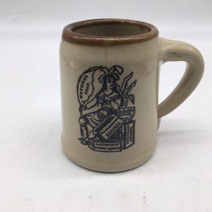 May include: Tan ceramic mug with a brown rim and handle. The mug features a black and white illustration of a woman holding a banner that reads "NATURAL LEAF" and a box labeled "HAVANA". The mug has a cylindrical shape.