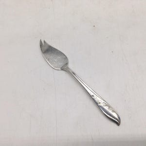 May include: A silver-toned metal cheese knife featuring a wide, pointed blade with a split tip and a slender handle decorated with an embossed floral pattern along the stem. The utensil has a polished finish and a curved, ergonomic design.