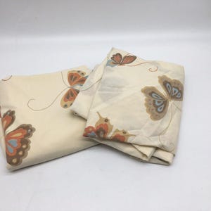 Puede incluir: Dos piezas de tela plegadas de color crema con un patrón repetitivo de mariposas en tonos naranja, marrón y azul claro. Las mariposas están representadas con líneas finas y curvas que muestran sus antenas y trayectorias de vuelo sobre el fondo.