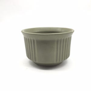 Könnte beinhalten: Ein salbeigrünes Keramik-Ramekin mit vertikalen Rippendetails. Die kleine, runde Schale hat einen leicht ausgestellten Rand und einen abgerundeten Boden. Das Ramekin ist zum Backen und Servieren einzelner Portionen bestimmt.
