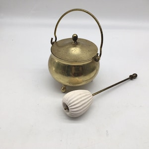 Puede incluir: Una pequeña olla de latón antigua con tapa y asa, apoyada sobre tres patas pequeñas. Un objeto a rayas blancas y grises con un mango largo de latón está al lado de la olla. La olla probablemente se usa para cocinar o servir.