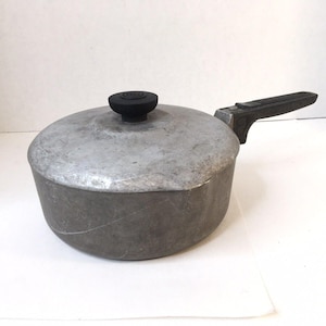 Può includere: Un pentolino rotondo in alluminio con finitura grigio opaco, dotato di coperchio coordinato con pomello circolare nero e un lungo manico nero resistente al calore che si estende lateralmente. La superficie metallica presenta segni di usura.