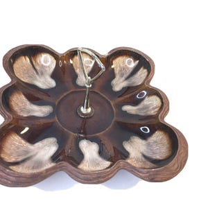 May include: A brown ceramic serving dish with a scalloped edge and a metal handle. The dish has a dark brown center with lighter brown and white accents in each of the eight sections. A decorative piece for serving appetizers or snacks.