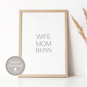 May include: A framed print with the text "Wife Mom Boss" in a light gray color on a white background. The frame is a light wood color. The print is sitting on a white surface with a vase of dried pampas grass.
