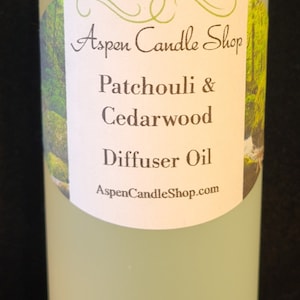 May include: A clear glass bottle with a white cap, labeled "Aspen Candle Shop Patchouli & Cedarwood Diffuser Oil AspenCandleShop.com". The label has a green background with a nature scene.