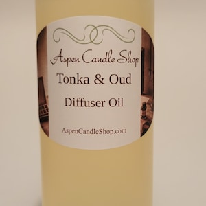 May include: A clear glass bottle with a white cap filled with a light yellow liquid. The label reads "Aspen Candle Shop Tonka & Oud Diffuser Oil AspenCandleShop.com"