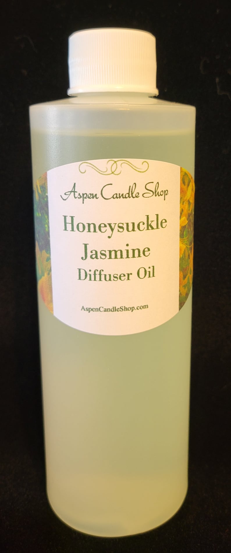 May include: A clear glass bottle with a white cap and a label that reads "Aspen Candle Shop Honeysuckle Jasmine Diffuser Oil AspenCandleShop.com". The label has a floral design with yellow and green flowers.
