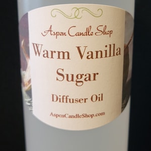 May include: A clear plastic bottle with a white cap, labeled "Aspen Candle Shop Warm Vanilla Sugar Diffuser Oil." The label is oval-shaped with brown text and a floral design. The bottle is filled with a clear liquid.