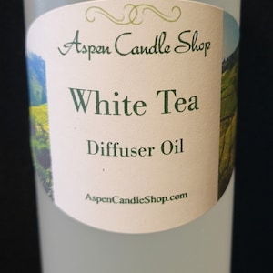 May include: A clear glass bottle with a white cap and a white label. The label has green text that reads "Aspen Candle Shop", "White Tea", and "Diffuser Oil". The label also has a website address: "AspenCandleShop.com".
