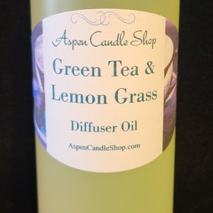 May include: A clear glass bottle with a white cap filled with yellow liquid. The label reads "Aspen Candle Shop Green Tea & Lemon Grass Diffuser Oil AspenCandleShop.com"