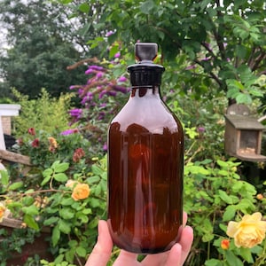 Puede incluir: Una botella de vidrio marrón oscuro con un tapón de vidrio negro. La botella tiene una forma redondeada y una boca ancha.