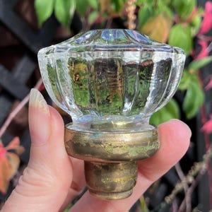 May include: Clear glass door knob with a ribbed design and a brass base. The knob has a rounded top and a cylindrical base. The brass base shows signs of age and wear. The background features green foliage.