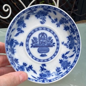 Puede incluir: Un platillo de cerámica blanco con un patrón floral azul y blanco. El centro del platillo presenta un diseño floral azul y blanco con una cesta.