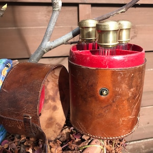 Puede incluir: Una caja de viaje de cuero marrón con forro rojo y cuatro viales de vidrio dorados en el interior. La caja tiene un cierre de metal y un detalle de costura decorativa.