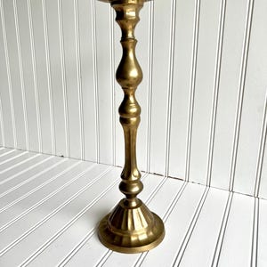 May include: A tall, brass-colored candlestick holder with a wide, shallow dish at the top. The stem has a bulbous design, and the base is round with a fluted edge. The candlestick holder is approximately 12 inches tall.