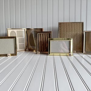 May include: A collection of vintage picture frames in various shapes and sizes. The frames are primarily gold-toned metal, with some featuring textured or patterned inserts. The frames are arranged on a white surface with a vertical stripe pattern.