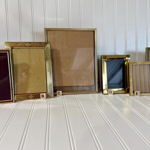May include: A collection of six vintage gold-toned picture frames of varying sizes. The frames have different designs, including rectangular and square shapes. Some frames have glass, while others have backing. The frames are arranged on a white surface.