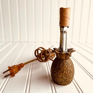 May include: A vintage pineapple-shaped lamp base in a warm, golden-brown hue. The lamp features a detailed, textured design and a brown electrical cord with a two-prong plug. The lamp is designed to hold a lightbulb and is ready for use.