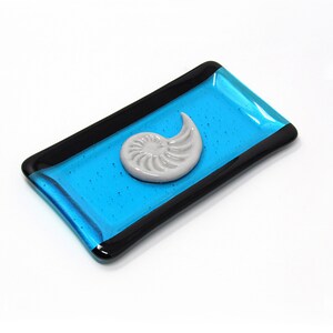 May include: A small rectangular dish made of glass with a black border. The dish is blue with a white nautilus shell design in the center.