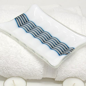 May include: A white glass soap dish with a wavy design of blue and gray stripes. The dish has a rectangular shape with slightly curved sides. It rests on a white towel, suggesting a bathroom or spa setting.