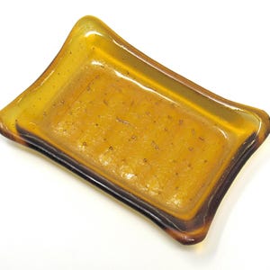 May include: A rectangular amber-colored glass dish with rounded corners. The dish has a slightly raised rim and a dark brown edge. The glass appears to have small bubbles within it, giving it a unique texture. The dish is likely used for holding small items.