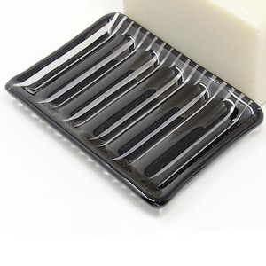 May include: A black glass soap dish with a rectangular shape and diagonal white stripes. The dish has raised ridges for drainage and is next to a white bar of soap. The soap dish is a bathroom accessory.