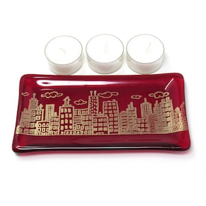 May include: A rectangular, ruby red glass candle holder with a gold cityscape design. Three white tea light candles sit above the holder. The cityscape features buildings, clouds, and architectural details.